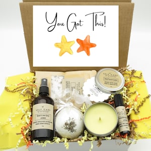May include: A gift box containing a room and linen spray, a bath bomb with dried flowers, a hand-poured candle in a tin, and an aromatherapy roller. The box lid features the text You Got This! with yellow and orange star illustrations.