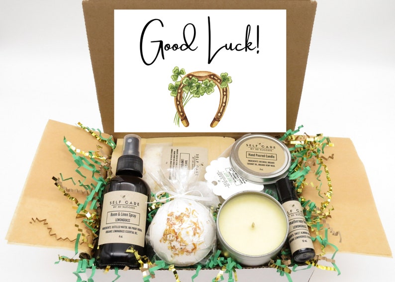 May include: A brown cardboard box filled with green and gold tissue paper. The box contains a variety of self-care items, including a candle, bath bomb, room spray, and essential oil. The box also includes a card with the text "Good Luck!" and a horseshoe with four-leaf clovers.