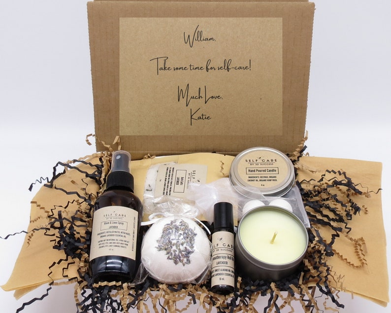 May include: A brown cardboard box filled with self-care items, including a lavender bath bomb, a bottle of body and linen spray, a candle, and a roll-on essential oil. The box is decorated with a handwritten note that says "William, Take some time for self-care! Much Love, Katie."