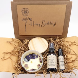 May include: A brown cardboard box filled with a self-care gift set. The box contains a candle with purple flowers and crystals, a small bottle of room spray, a roll-on essential oil, and a wooden lid. The box is decorated with a card that says "Happy Birthday!"