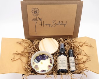May include: A brown cardboard box filled with a self-care gift set. The box contains a candle with purple flowers and crystals, a small bottle of room spray, a roll-on essential oil, and a wooden lid. The box is decorated with a card that says "Happy Birthday!"