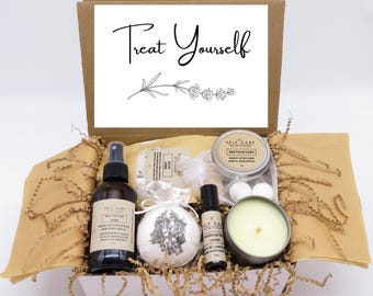 May include: A cardboard gift box containing a lavender-scented self-care set. Items include a brown glass spray bottle, a round bath bomb with dried lavender, a small roller bottle, and a metal tin candle. The box lid features the text Treat Yourself.