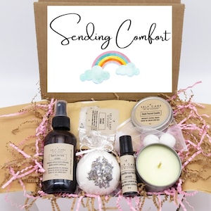 May include: A cardboard gift box containing a room and linen spray, a lavender bath bomb, a roller bottle, and a candle in a tin. The box lid features the text Sending Comfort and a rainbow graphic. The items are nestled in pink and tan paper filler.
