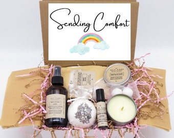 May include: A cardboard gift box containing a room and linen spray, a lavender bath bomb, a roller bottle, and a candle in a tin. The box lid features the text Sending Comfort and a rainbow graphic. The items are nestled in pink and tan paper filler.