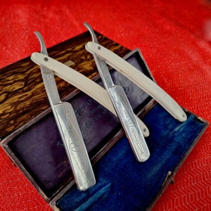 Puede incluir: Dos navajas rectas antiguas con mangos color marfil y hojas plateadas, marcadas "Hamburg Ring". Las navajas se exhiben en una caja de madera abierta con interior de terciopelo azul, sobre un fondo rojo.