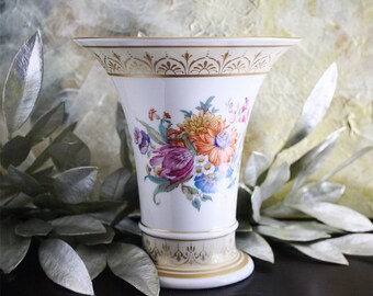Puede incluir: Un jarrón de porcelana blanca con un borde dorado y un diseño floral. El jarrón tiene una boca ancha y una base estrecha. Las flores están pintadas en tonos de rosa, naranja, amarillo y azul.