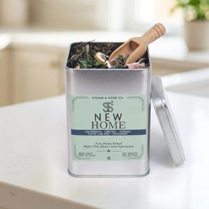 Puede incluir: Una lata rectangular de metal plateado llena de hierbas secas y una pequeña cuchara de madera. La etiqueta dice Steam & Sage Co. New Home, enumerando ingredientes como hierba de limón, cáscara de lima, pandan, cáscara de lima kaffir y menta.