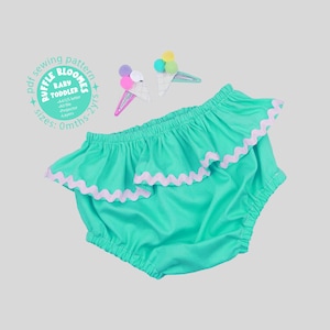 May include: A bright turquoise ruffled diaper cover with light purple rickrack trim, accompanied by two ice cream cone hair clips featuring colorful pom-poms. Text on the image reads: Ruffle Bloomes Baby Toddler sewing pattern sizes 0mths-2yrs.