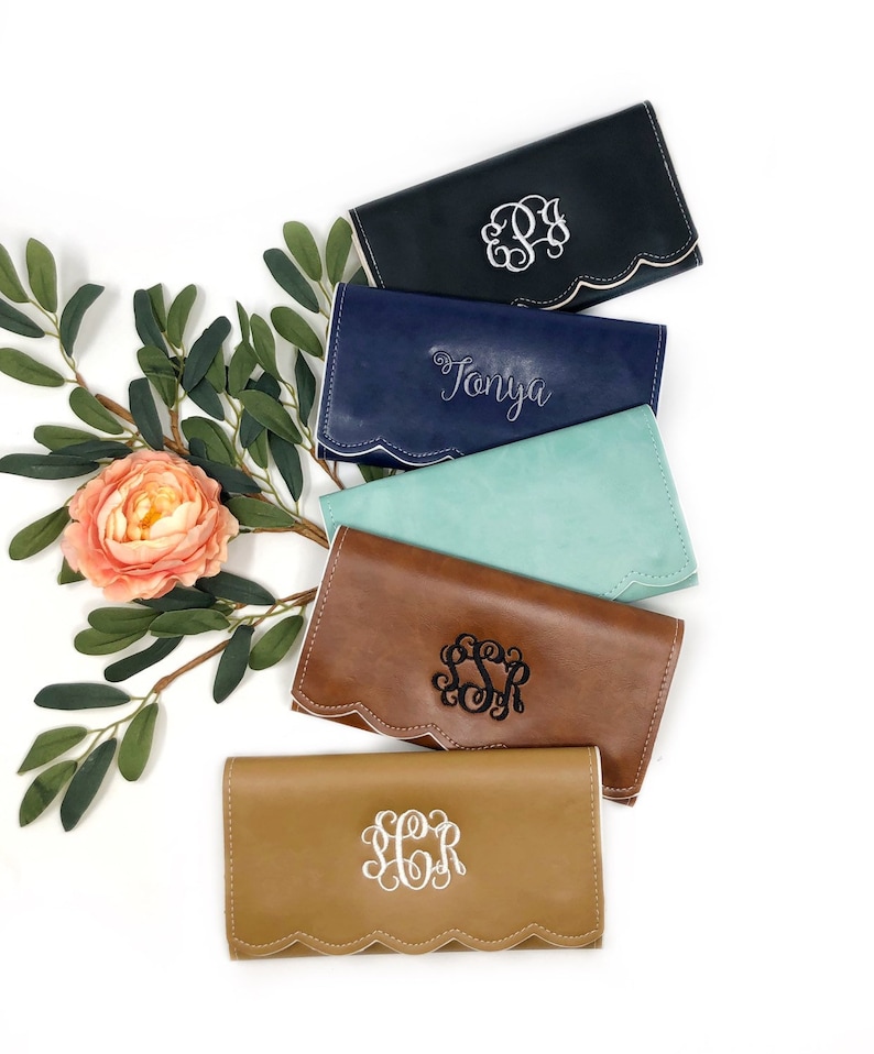 Puede incluir: Cinco carteras de cuero de diferentes colores con bordes festoneados. Las carteras est&aacute;n monogramadas con iniciales o un nombre. Las carteras est&aacute;n apiladas, con la cartera superior de color azul marino, seguida de azul marino, turquesa, marr&oacute;n y beige.