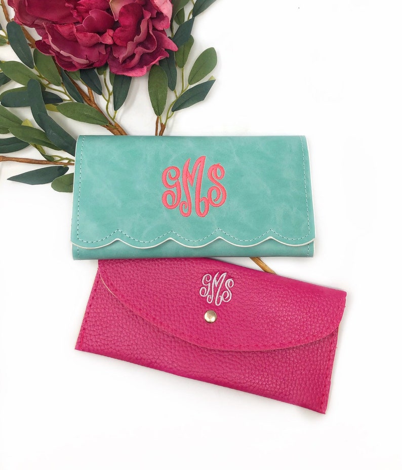 Puede incluir: Dos carteras monogramadas, una turquesa con bordes blancos festoneados y la otra rosa intenso. Ambas carteras presentan el monograma "SMS" en una fuente de letra blanca.