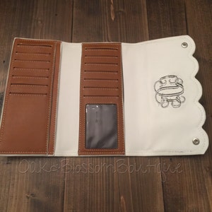 Puede incluir: Cartera de cuero blanco de tres pliegues con interior de cuero marr&oacute;n y borde festoneado. La cartera tiene m&uacute;ltiples ranuras para tarjetas, una ventana de identificaci&oacute;n transparente y una inicial bordada.