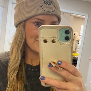 May include: A beige beanie with a green and black embroidered Grinch face. The beanie is folded down and has a ribbed texture.