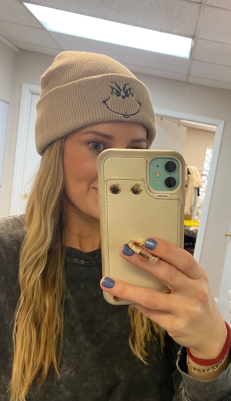 May include: A beige beanie with a green and black embroidered Grinch face. The beanie is folded down and has a ribbed texture.