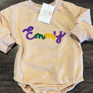 May include: A light peach-colored baby romper with long sleeves and ribbed cuffs. The name "Emmy" is embroidered in purple, yellow, and green thread across the chest. A tag with the brand name is attached to the neckline.