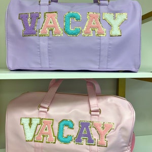 Pu&ograve; includere: Due borsoni da viaggio con la parola "VACAY" scritta in lettere colorate e pelose. La borsa in alto &egrave; lavanda e la borsa in basso &egrave; rosa.
