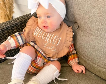 以下が含まれることがあります： 白いリボンカチューシャ、名前「WRONGLEY KATE」の茶色のよだれかけ、チェック柄のワンピースを着た赤ちゃん。赤ちゃんは白いニーハイソックスとリボン付きの白い靴も履いています。赤ちゃんは灰色のクッションに座っています。