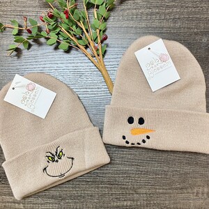 May include: Two beige knit beanies with embroidered designs. One beanie features a snowman face with black eyes, a carrot nose, and a black smile. The other beanie features a green Grinch face with black eyes, a black mouth, and a black nose.