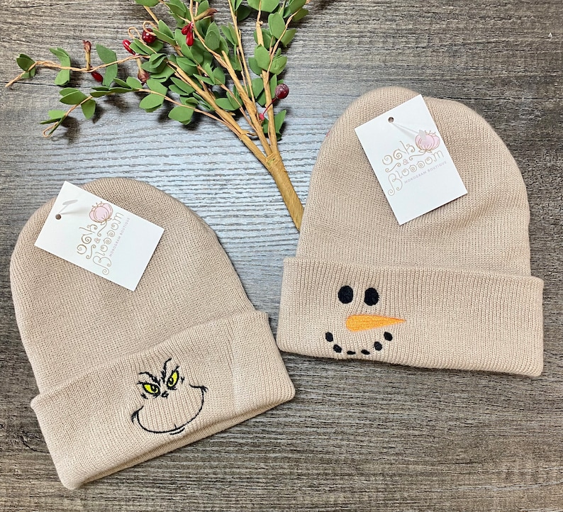 May include: Two beige knit beanies with embroidered designs. One beanie features a snowman face with black eyes, a carrot nose, and a black smile. The other beanie features a green Grinch face with black eyes, a black mouth, and a black nose.