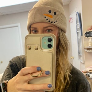 May include: A beige knit beanie with a snowman design featuring black eyes, a black mouth, and an orange carrot nose.