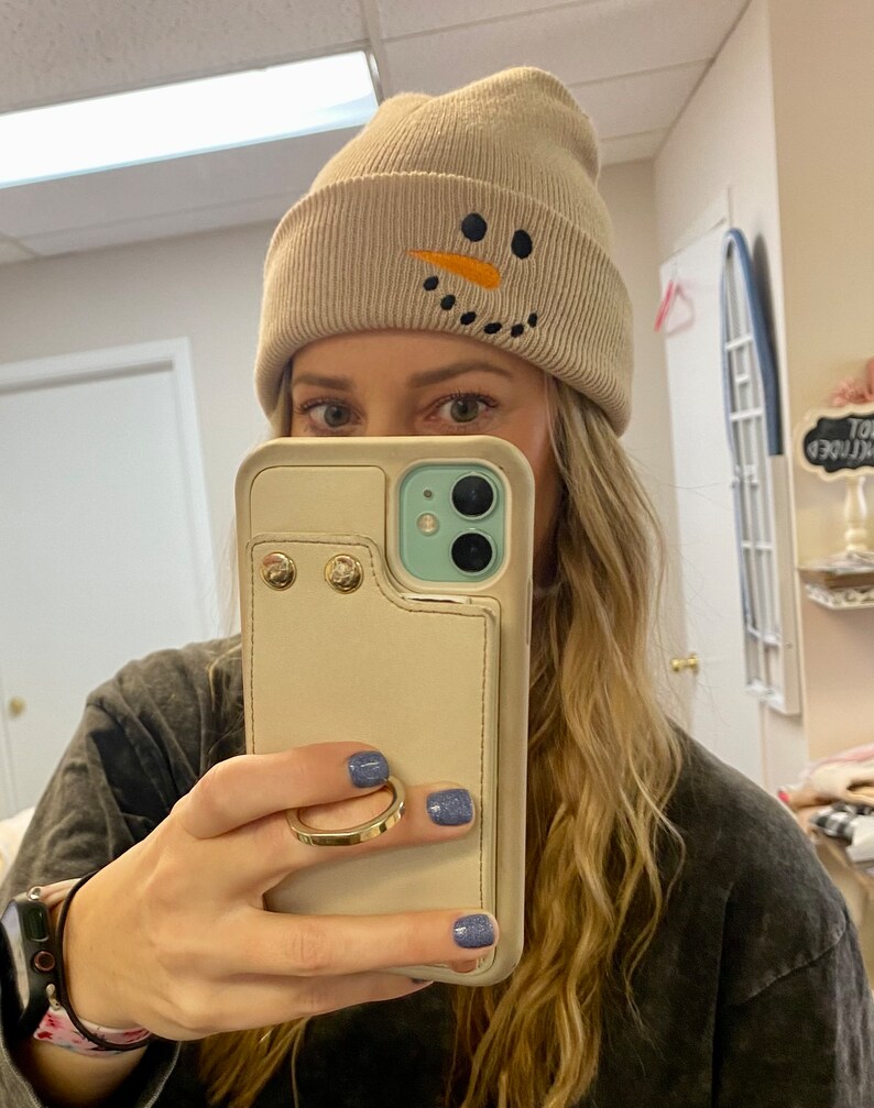 May include: A beige knit beanie with a snowman design featuring black eyes, a black mouth, and an orange carrot nose.
