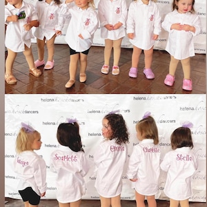 Puede incluir: Ocho niñas pequeñas con camisas blancas abotonadas con nombres personalizados bordados en la espalda. Las niñas están de pie en dos filas, mirando a la cámara. El fondo es una pared blanca con el texto "helena hosch dancers" impreso en ella.