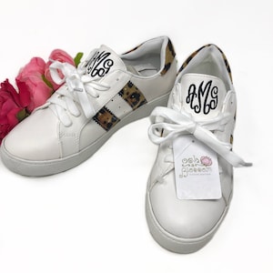 May include: White leather sneakers with a leopard print stripe and black monogram "AMS" on the side. The sneakers are tied with white laces and have a white rubber sole.
