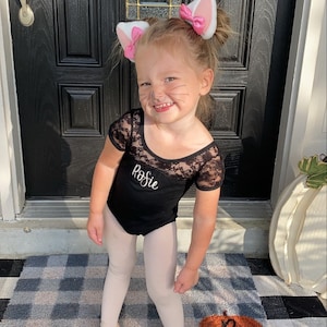 May include: A young girl dressed as a cat for Halloween. She is wearing a black leotard with lace detailing and the name "Rosie" embroidered on the front. She is also wearing pink cat ears and has cat whiskers drawn on her face. She is standing on a black and white checkered rug.