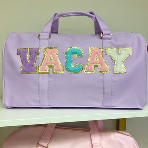 Op de afbeelding: Paarse en roze reistas met het woord "VACAY" in kleurrijke letters met gouden glitteraccenten.