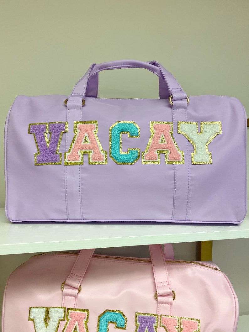 Pu&ograve; includere: Borsa da viaggio viola e rosa con la parola "VACAY" in lettere colorate con accenti di glitter dorati.
