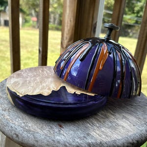 May include: A handcrafted wooden box with a lid. The box is painted a deep purple with black and orange drips. The lid has a small, dark metal handle. The interior of the box is a light, natural wood color.