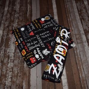 May include: Two black fabric wristlets with white and yellow lettering. One wristlet has the word "ZADDY" written in white letters. The other wristlet has a black and white pattern with the words "100% Happy", "Mickey", "Love", and "Toy".