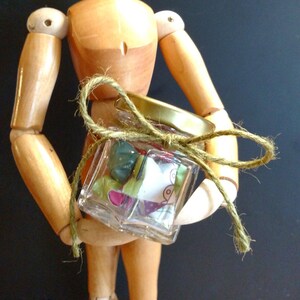 Peut inclure: Un mannequin en bois d'artiste tenant un petit bocal en verre avec un nœud en ficelle brune. Le bocal est rempli de chutes de tissu colorées et à motifs.
