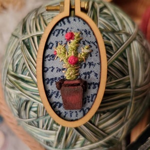 Op de afbeelding: Een kleine, geborduurde cactus in een bruine pot, ingelijst in een houten ring. De cactus is groen met rode bloemen en is geplaatst tegen een blauwe achtergrond.