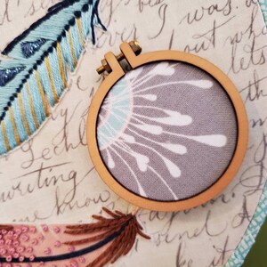 以下が含まれることがあります： グレーの布地に白い花柄が描かれた、小さな木製の刺繍枠のクローズアップ。枠は、手書きの文字と青と緑の刺繍された羽根が描かれた布の上に置かれています。
