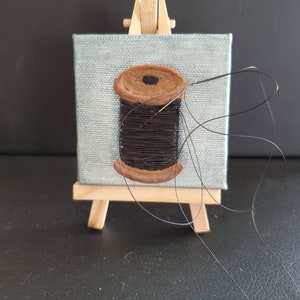 Op de afbeelding: Een klein schilderij op canvas van een bruine spoel met zwarte draad, met een naald en draad die uit de spoel komen. Het schilderij staat op een klein houten ezel.