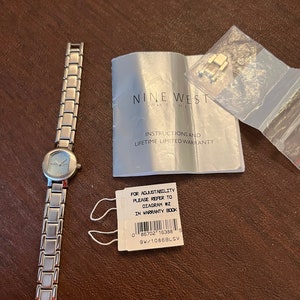 May include: A silver-toned Nine West watch with a white face and silver hands. The watch comes with instructions and a lifetime limited warranty. The packaging includes a small plastic bag with a spare watch link.