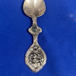 Pode incluir: Uma colher de prata com desenhos florais intrincados e um detalhe de pedra turquesa. O verso da colher apresenta uma imagem detalhada de um dragão.