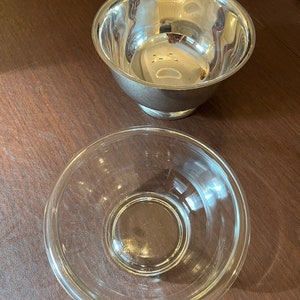 Könnte beinhalten: Zwei Schalen, eine silberne und eine aus klarem Glas, stehen auf einer braunen Holzoberfläche. Die silberne Schale hat eine abgerundete Form und eine glänzende Oberfläche. Die Glasschale ist klar und hat einen leicht breiteren Rand.