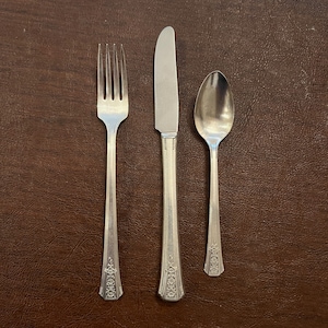 May include: A set of three silver-plated cutlery items, comprising a fork, knife, and spoon. The cutlery features an ornate design with floral patterns.