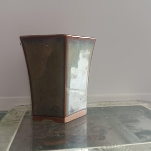 May include: A hexagonal ceramic planter with a green and brown glaze. The planter has a wide base and a narrow opening.