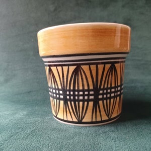 Pode incluir: Um pequeno vaso de cerâmica com um padrão geométrico em preto e branco sobre um fundo castanho claro. O vaso tem uma forma arredondada e uma borda ligeiramente alargada.