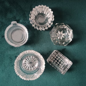 Puede incluir: Vista aérea de cinco recipientes de vidrio tallado transparente. Los recipientes tienen varias formas, incluyendo redonda, hexagonal y rectangular. El vidrio presenta patrones y texturas intrincados, con diseños de estrella y geométricos. Están dispuestos sobre una superficie verde oscuro.