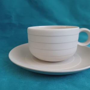 May include: White ceramic teacup and saucer set. The cup has horizontal grey lines and a rounded handle. The saucer is round with concentric circles. The set is on a teal background.