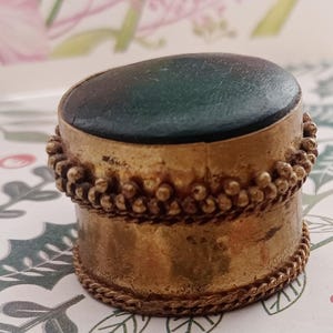 May include: A small cylindrical container with a dark green circular lid and a metallic gold-toned body. The exterior features decorative beaded and twisted wire trim around the top and bottom edges, creating a textured, antique-style finish.