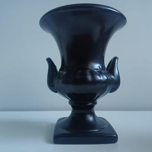 May include: A black ceramic urn with a square base and two handles on the sides. The urn is a classic design with a smooth, glossy finish.