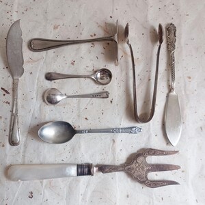 Puede incluir: Una variedad de cubiertos antiguos de plata, que incluyen un cuchillo de mantequilla, cucharas, pinzas, un tenedor de servir y un cuchillo de servir. Los cubiertos plateados están dispuestos sobre una superficie texturizada blanquecina.