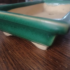 Peut inclure: Un pot rectangulaire en céramique verte avec un intérieur brun clair et quatre petits pieds. Le pot est en céramique émaillée et a une finition lisse.