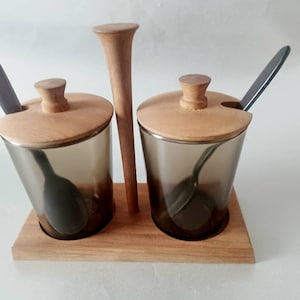 Peut inclure: Un présentoir à épices en bois avec deux récipients en verre, chacun avec un couvercle et une cuillère en bois. Un ustensile en bois se trouve entre les récipients. Les récipients en verre sont gris fumé, et les cuillères sont gris foncé.