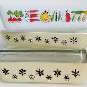 May include: A collection of vintage Pyrex baking dishes. The top dish is white with a colourful vegetable design. The middle dish is cream-coloured with black snowflakes. The bottom dish is cream-coloured with black snowflakes and a glass lid.