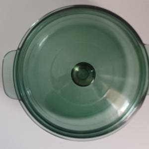 Puede incluir: Vista cenital de una fuente redonda de vidrio verde con su tapa a juego. La fuente tiene dos asas y un pomo de vidrio transparente en la tapa. La fuente está hecha de vidrio transparente y resistente al calor.